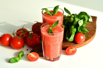 Two glasses of fresh organic tomato juice decorated with raw tomatoes and green leaves of basil on light background.Healthy diet nutrition concept.