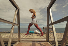 Attractive Young Blonde Woman With Crimped Hair, Wearing A Striped Dress And Long Red Boots, Walking On The Dock Of A Lobster Fisherman's Cove
