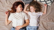 Relaxed Brunette Mother And Curly Haired Preschooler Daughter Lie On Large Grey Bed And Kiss Near Black White Pillows Close Upper View