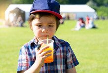High Key Portrait Of Kid Boy Drinking Orange Slush From Plastic Glass In Hot Sunny Day,Thirsty Child Drinking Frozen Orange Slush While Waking At The Park, Summer Drink For The Kids