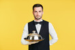 © GVS - Handsome elegant waiter holding tray and cloche ready to serve on yellow background