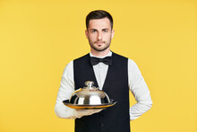 Handsome Elegant Waiter Holding Tray And Cloche Ready To Serve On Yellow Background