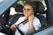 © Sergio - Mature smiling woman with glasses sitting in car behind the wheel and wearing seat belt. Senior woman driver buckles up and looks in side mirror of car. Active pensioner, road trip, driving safety.