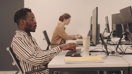 Side-view shot of serious African-American IT student or programmer typing on computer sitting by desk with female coworker in modern room