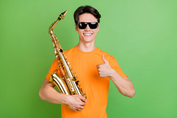 Sticker - Photo of positive happy young man make thumb up wear sunlgass hold hand sax isolated on green color background