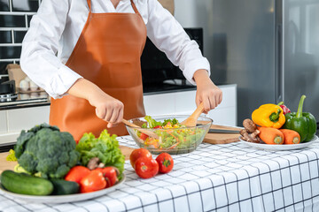 Wall Mural - Asian housewife wearing apron and using ladle to mixing vegetable salad in bowl while standing preparing