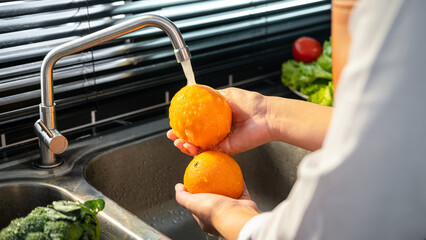 Wall Mural - Hands of asian housewife washing orange and vegetables with water in sink while wearing apron