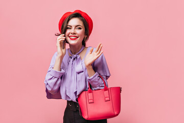 Wall Mural - Portrait of girl in lilac blouse and red beret on pink background. Woman holding small bag and talking on phone