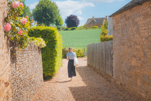 A Young Blonde Female Tourist Explores A Quaint Country Lane In The Rural English Countryside Village Of Chipping Campde, On A Summer Day In The Cotswolds, Gloucestershire, UK.