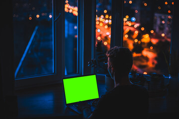 Wall Mural - A laptop with an empty green screen on a wooden table at night with a view of the city with colored lights. A place for text, the concept of programming and development