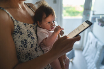 Adult woman mother holding three months old baby while using looking mobile phone for internet browsing or social network texting in room at home domestic life motherhood and parenting busy concept