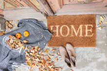 Doormat With Plaid, Shoes And Leaves Near Entrance Of House On Autumn Day