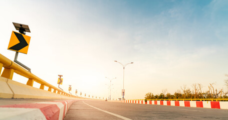 Wall Mural - Curve concrete road with curve traffic sign beside the sea at sunset time. Solar panel energy on yellow curve traffic sign. Road trip on summer vacation. Blur car driving. Summer travel by car.