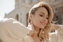 Cheerful Blonde Curly Woman In Pearl Necklace And White Elegant Blouse Smiles Sincerely, Looks Down And Poses Outside.