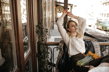 Wall Mural - Attractive curly blonde lady in pearl necklace and white blouse rises arms and sits in cozy street cafe with yellow bouquet.