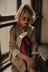 Wall Mural - Serious blonde woman with ponytail and bright lips in red sunglasses and beige trench coat sits on windowsill and looks into camera near windows.