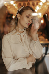 Wall Mural - Charming blonde woman in stylish white shirt and pearl necklace gently touches hair. Young lady in light blouse poses in restaurant.