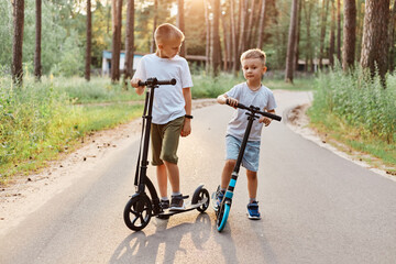 Outdoor shot of two brother wearing casual clothing riding scooters in summer park, spending time happily, having fun together in active way, happy childhood.