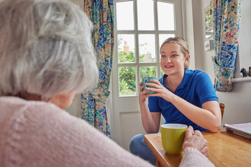 Wall Mural - Female Home Help Having Cup Of Tea With Lonely Senior Woman In Kitchen