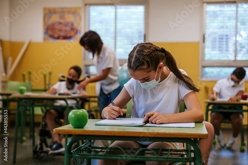 A girl student, wearing a protective mask, sits at a desk writing in her notebook in the background a teacher helps a disabled student sitting in a wheelchair. Back to school concept
