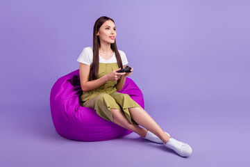 Sticker - Full length photo of young focused happy woman look empty space play game sit chair isolated on violet color background