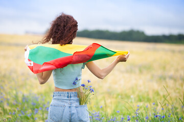 Woman holding flag of Lithuania in a rye field with blue cornflowers. Back view. Lithuanian Flag Day. Independence restoration Day. Travel and love Lithuania concept. Selective focus.