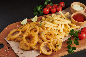 Wall Mural - Homemade baked potato fries with mayonnaise, Tomato sauce and rosemary on wooden board. fast food products : onion rings, french fries on cutting board, on black stone background, unhealthy food.