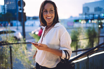 Cheerful beautiful woman in trendy wear and eyeglasses for vision protection laughing from funny social media content, pretty female blogger 30s holding mobile phone and rejoicing outdoors