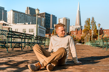 A young man sits on a wooden floor on a pier in the morning with a view of downtown and the Transamerica Tower in San Francisco