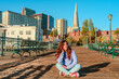 © KseniaJoyg - A young woman in jeans sits on a wooden floor on a pier in the morning with a view of downtown and the Transamerica Tower in San Francisco