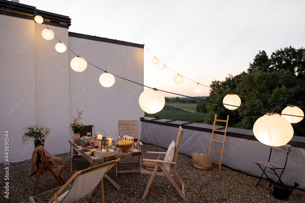 Beautifully decorated roof terrace in natural boho style with a dining table full of food at summer time. Festive dinner for a small group of friends at dusk outdoors - obrazy, fototapety, plakaty 