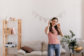 Wall Mural - Model has cool time in her cozy apartment surrounded by favorite sofa and bookshelves. Girl in tight jeans and tee listening to music in headphones and dancing