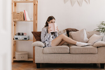 Wall Mural - Cute woman in pajamas and socks is resting on sofa and covers her face with book. Pretty young lady in home outfit holds pink notebook and sits on beige couch