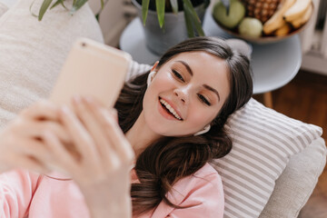 Wall Mural - Woman in great mood takes selfie in living room. Girl listening to music and lying on couch at home. Attractive woman smiling and holding smartphone