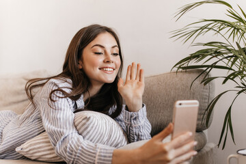 Wall Mural - Cheerful girl in striped shirt is lying on couch and talking via video in phone. Woman in pajamas waves her hand while having call with friend