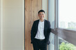 © Liubomir - Freelancer Asian businessman in business suit looking at camera near the window of a modern office center