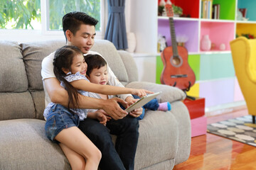happy asian dad teaching his daughter to use or play tablet on grey sofa in living room with laughing and smiling face (lifestyle with technology concept)