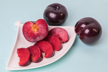 On a white saucer there is a large plum cut into wedges, next to it there are two more plums. The pulp of red berries. Close-up.