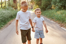 Two Little Boys Wearing White T Shirts And Shorts Going Together And Holding Hands In Summer Park, Brothers Walking Outdoor, Expressing Positive Emotions.
