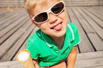 Happy funny little boy in stylish sunglasses looking at camera and showing tongue while eating tasty ice cream in summer park