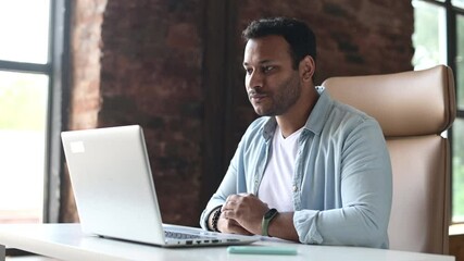 Wall Mural - Young indian male employee is using video connection for virtual meeting with co-workers, eastern guy sits in front of the laptop in contemporary office, greeting and waving into webcam