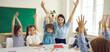 © Studio Romantic - Happy children and educator raising hands up sitting at table in modern classroom. Website header with group portrait of cheerful school teacher and first grade students having fun activities in class