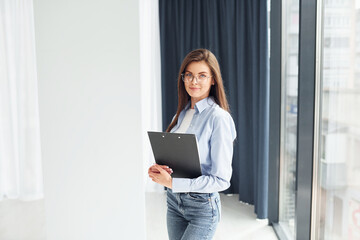 Young woman in glasses, formal clothes and with notepad standing indoors