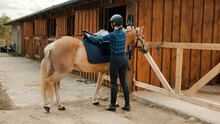Female Jockey Saddling Up Her Light Brown Horse Outside The Stable. Placing A Leather Saddle On The Back Of Her Horse. The Girl Wearing A Helmet And Gloves. Preparing Her Horse For The Competition.