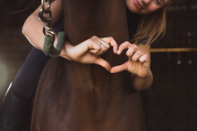 Horsewoman Posing With Her Seal Brown Horse In The Stable. Girl Making A Heart With Her Fingers. Expressing Her Love For The Stallion. Bonding Between Human Beings And Horses Concept.