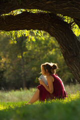 sunny portrait of a beautiful girl sitting on green glade under an arch of tree branches with book, woman reading novel on nature, concept hobby and lifestyle