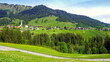 Weg im Kleinwalsertal mit Dorf Mittelberg umrahmt von grünen Wiesen und Wald unter blauem Himmel
