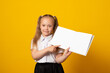 Education and school concept. Smiling little schoolgirl with book. Copy space. Little kid looking at mockup poster and standing on yellow background.