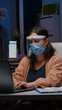 © DC Studio - Workaholic tired businesswoman with face mask and visor against covid19 working in company office typing financial statistics on laptop. Manager remains alone in company room after collegue left