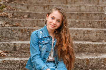 Wall Mural - A cute little girl in a denim jacket is sitting on the steps covered with leaves